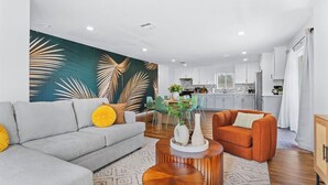 A modern living room with a grey sofa, orange armchair, and a wooden coffee table. A kitchen is visible in the background with white cabinets and a dining area. - Wonderful Home near DTLA/Crypto Arena/Universal Studio (Los Angeles)