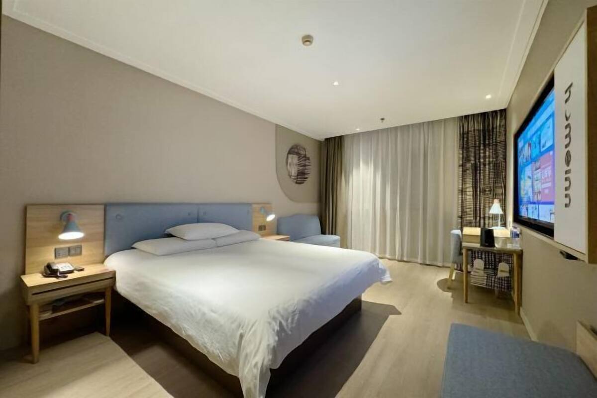 A hotel room with a large bed, bedside tables, a desk, a chair, a TV, and curtains.