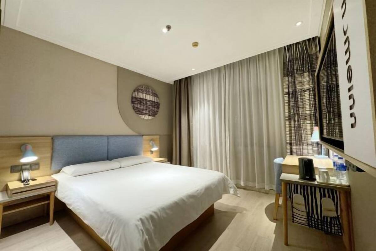 A modern hotel room with a large bed, bedside tables, a desk with a chair, a TV, and curtains.
