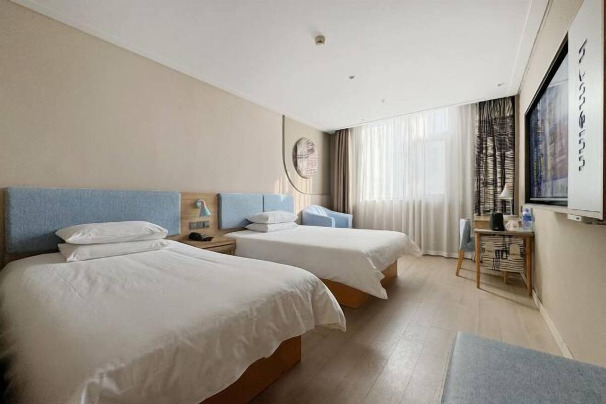 A hotel room with two beds, a desk, a chair, a lamp, and a window with curtains.