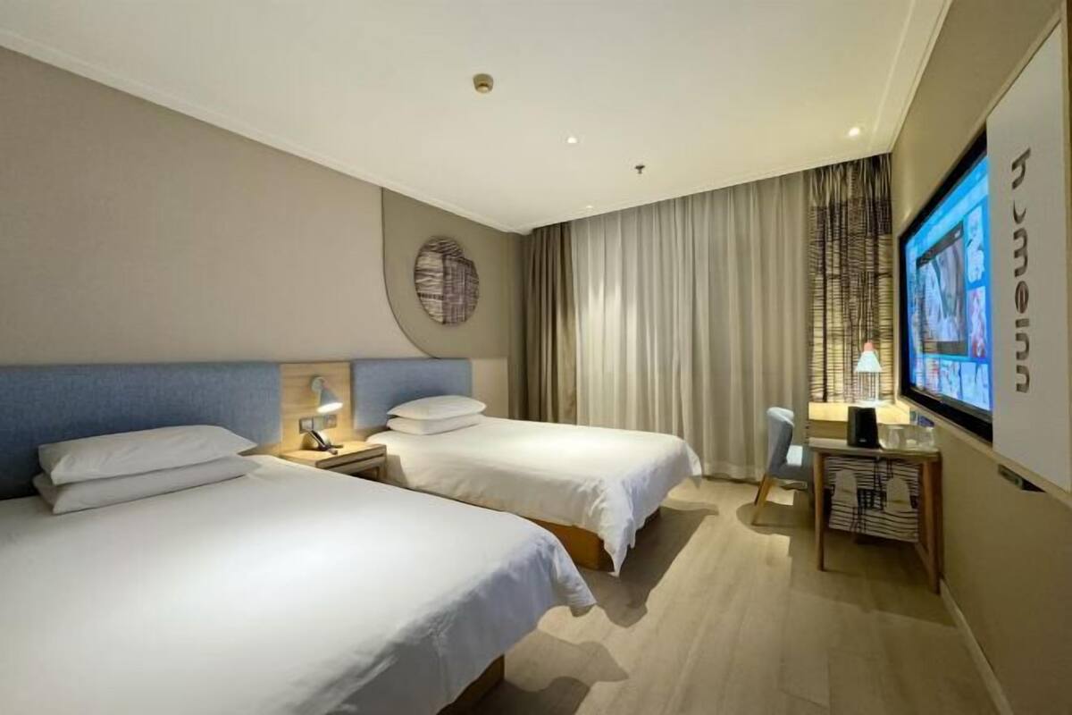 A hotel room with two beds, a desk, a TV, and a large window with curtains.