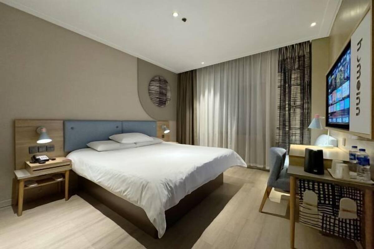 A modern hotel room with a large bed, a desk with a chair, a TV, and a wall-mounted screen displaying content.