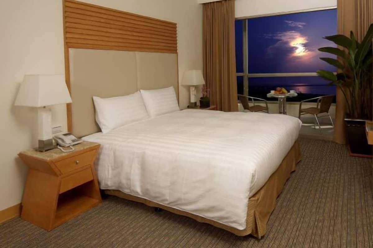 Una habitación de hotel con una cama grande, una mesita de noche con teléfono, una pequeña mesa con lámpara y vistas a la puesta de sol sobre el agua.