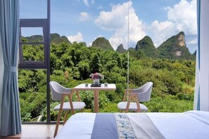 A bedroom with a large window, a bed, two chairs, a small table, and a view of a mountainous landscape. - Yangshuo Liyin Homestay (Yangshuo)