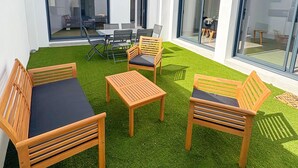 A modern outdoor patio with wooden furniture and artificial grass, featuring large sliding glass doors and a clear view of the surrounding area. - Cosy, spacious house near Puy du Fou (La Boissière-de-Montaigu)