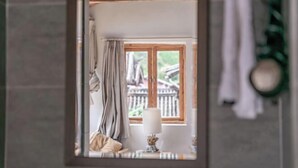Um banheiro com espelho refletindo uma janela com cortinas, um porta-toalhas e um abajur.