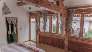 Quarto com cama, vigas de madeira, janelas amplas com cortinas e vista para o exterior.