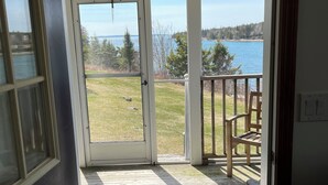 A wooden deck with a glass door leading to a grassy area with a view of a lake and trees. - Charming 3-bedroom waterfront cottage in Tremont with spectacular sunsets. (Tremont)