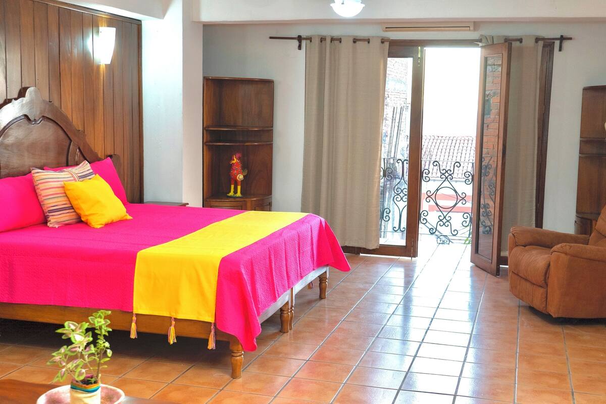 Une chambre d’hôtel avec un grand lit, une tête de lit en bois, un fauteuil brun et un balcon agrémenté d’une grille en fer forgé.