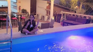 Zona de piscina con una persona sentada junto a la piscina, rodeada de mobiliario exterior y un patio cubierto con luces colgantes.