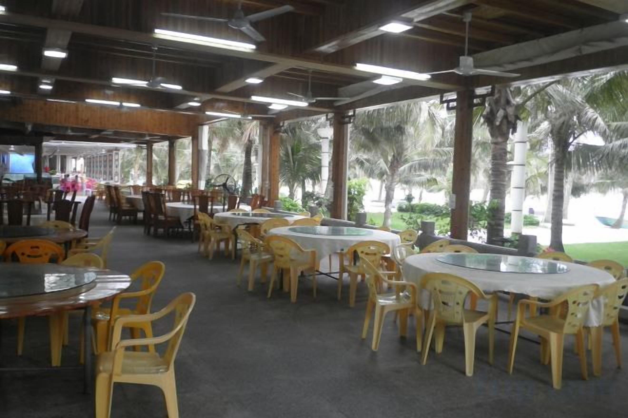 An indoor dining area with round tables and chairs, a covered roof, and a view of palm trees.
