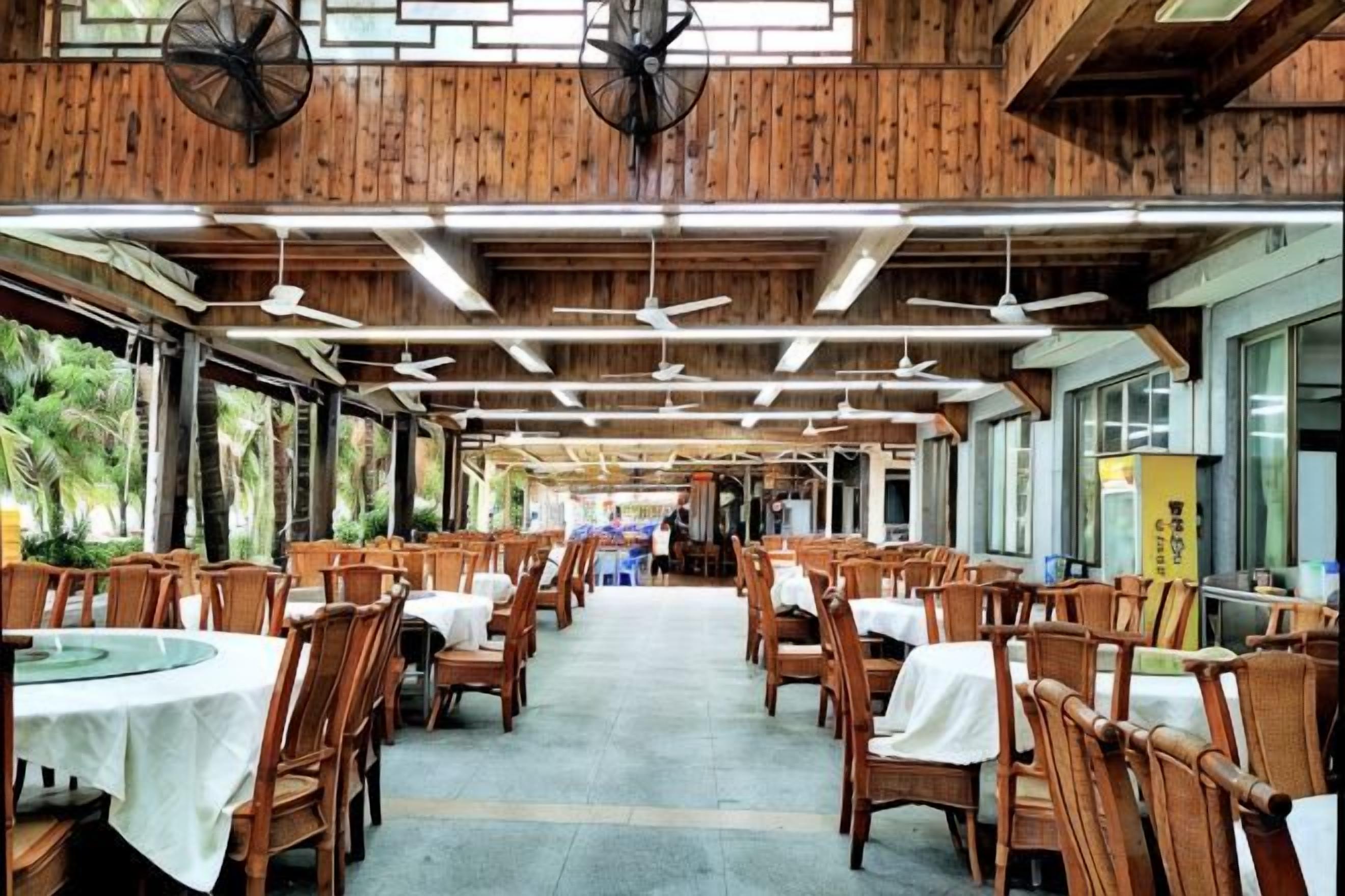 Un comedor interior con paredes y techo de madera, ventiladores de techo, mesas con manteles blancos y sillas.