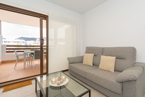 A living room with a grey sofa, two pillows, a glass coffee table with decorative items, and a sliding glass door leading to an outdoor balcony. - Apartamento Narcisos (Mar de Pupi) (San Juan de los terreros)