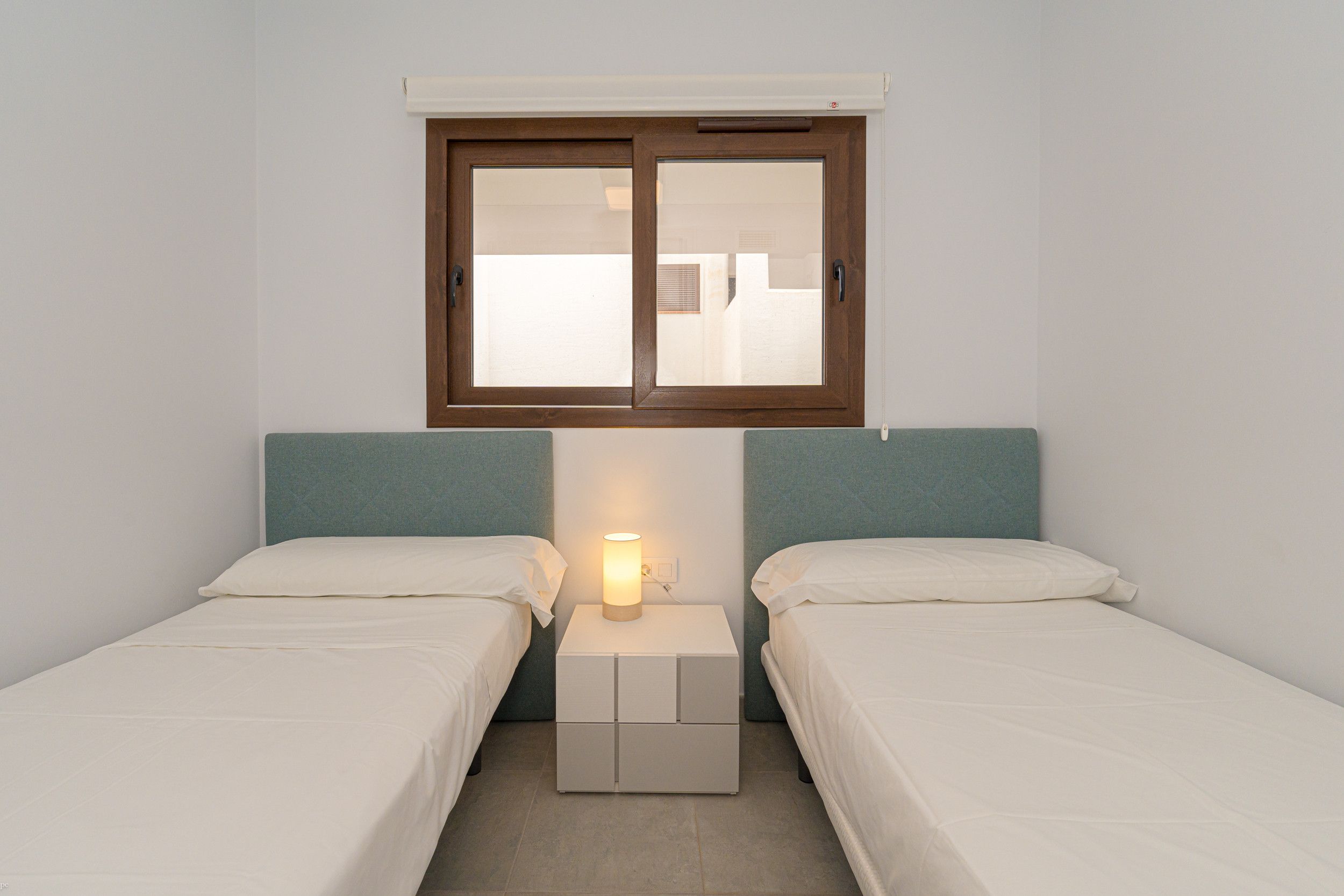 A small, clean room with two single beds, a nightstand with a lamp, and a window above the beds.