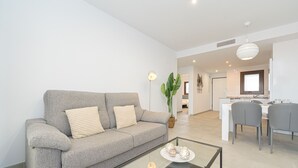 Ein modernes Wohnzimmer mit einem grauen Sofa, einem Glastisch und einem Essbereich mit grauen Stühlen und einem weißen Tisch.