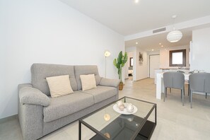 A modern living room with a grey sofa, a glass coffee table, and a dining area with grey chairs and a white table. - Apartamento Narcisos (Mar de Pupi) (San Juan de los terreros)