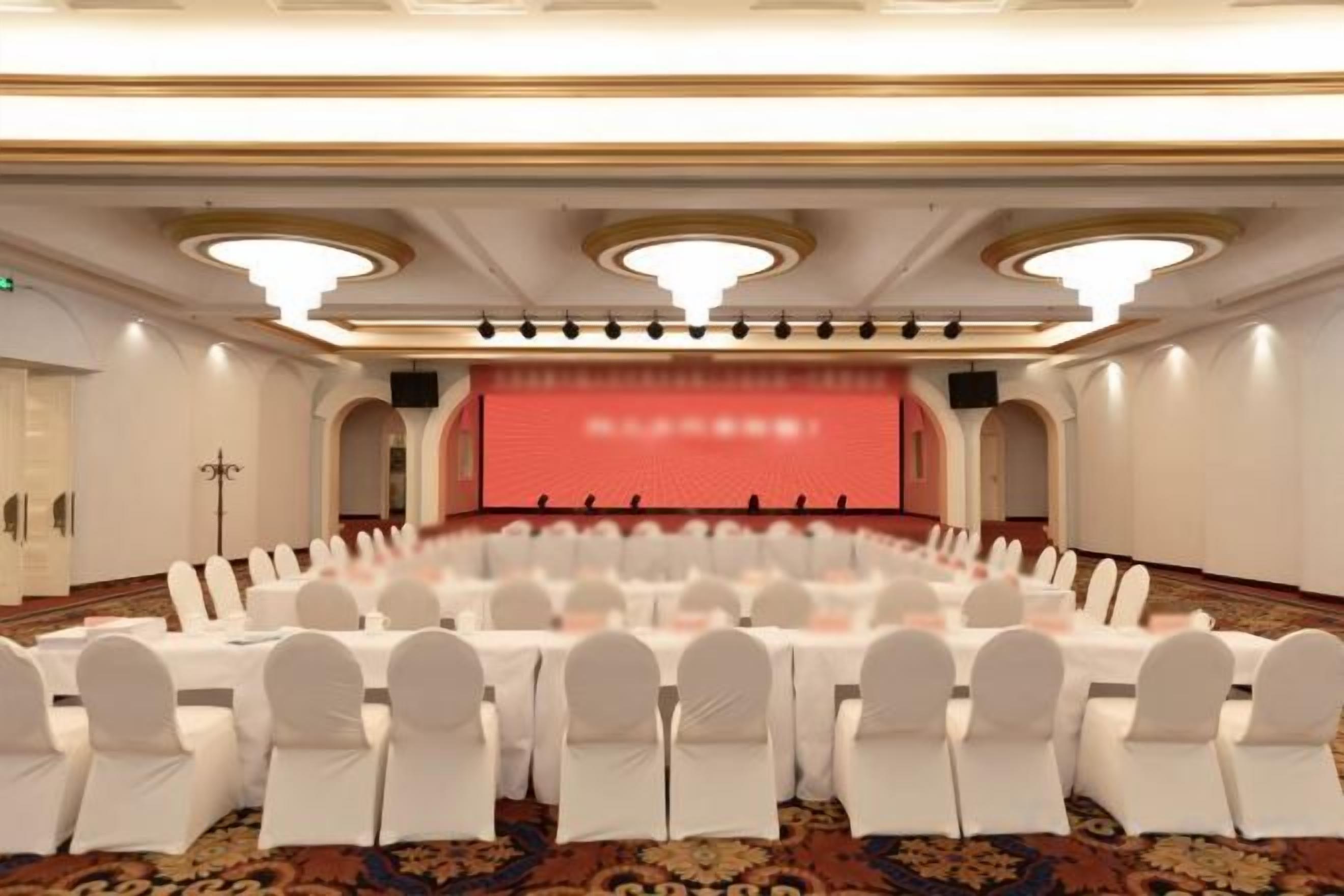 A conference room with round tables and white chairs, a stage with a red backdrop, and a carpeted floor.