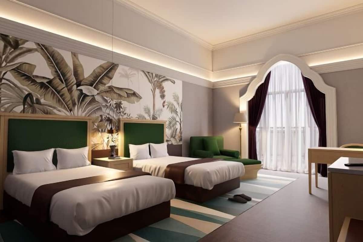 A hotel room with two beds, a large tropical mural, a green accent wall, a desk, and a window with curtains.