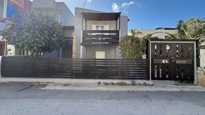 A modern two-story building with a gated entrance, a sign for a speed wheel car rental, and a sign for Dom Suites. - DOM SUITES (Daratsos)