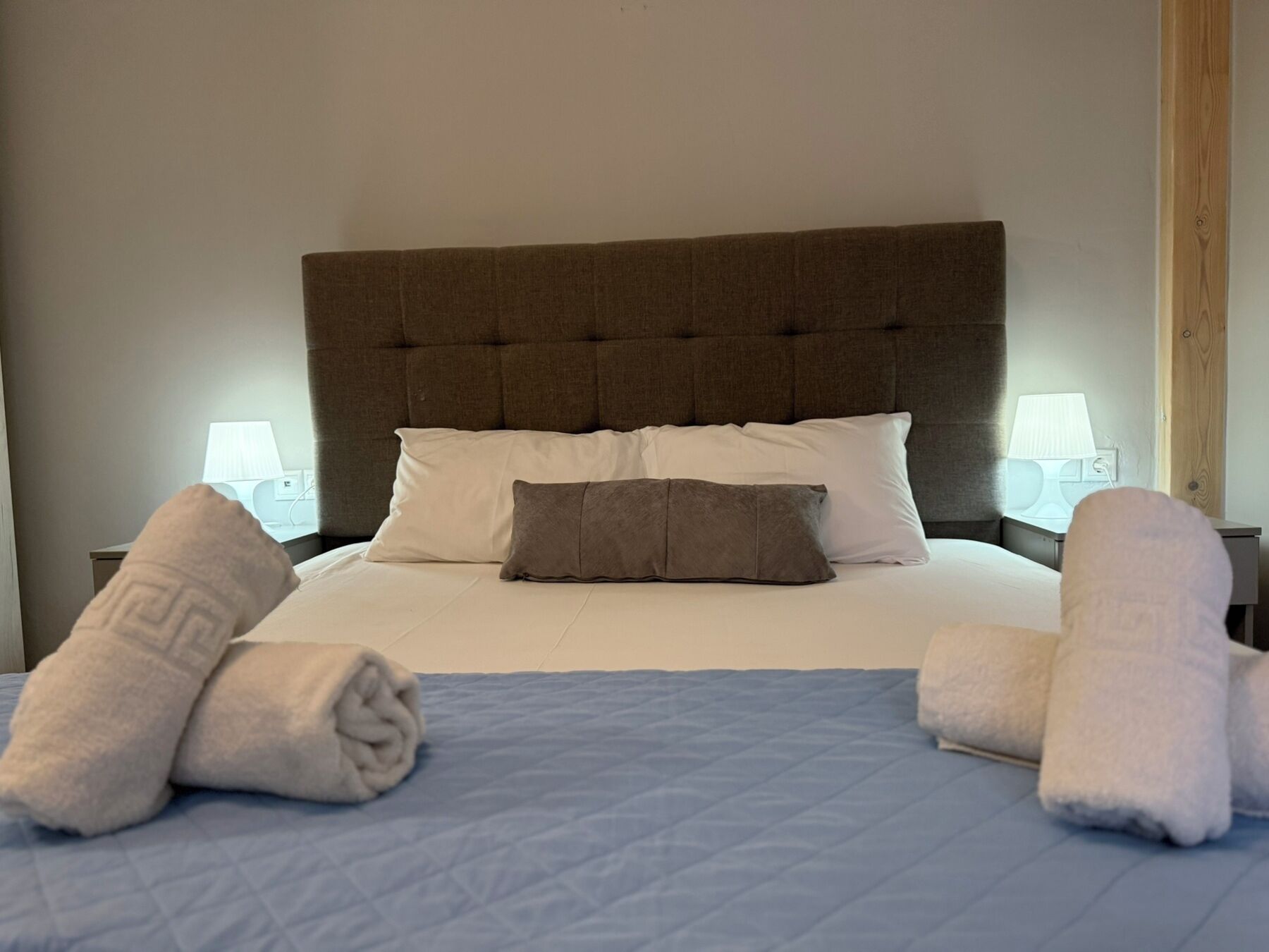 A neatly made bed with a brown headboard, white and gray pillows, and two rolled towels on either side.