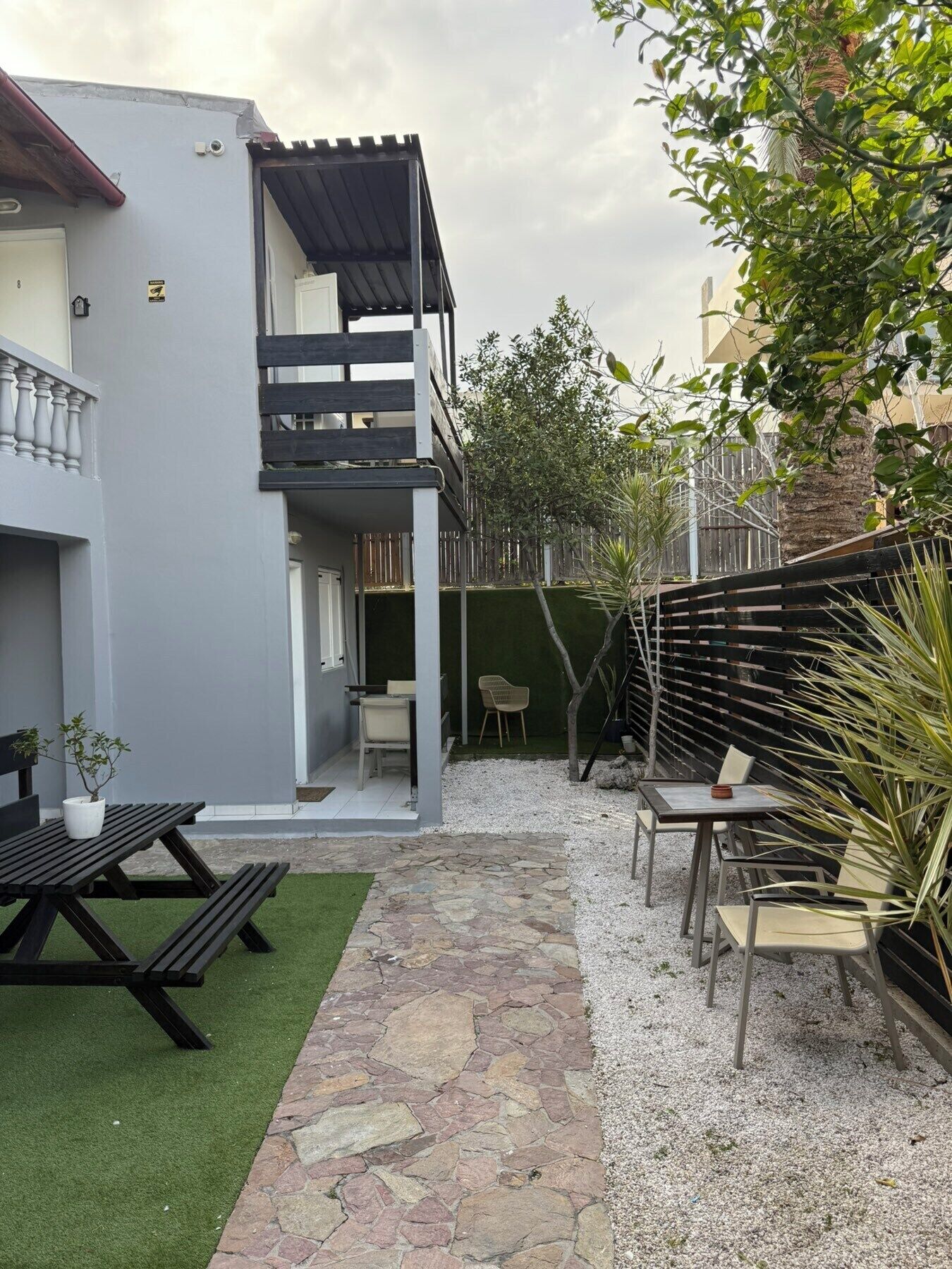 A two-story building with a balcony, a patio area with a table and chairs, and a small garden with a tree and plants.