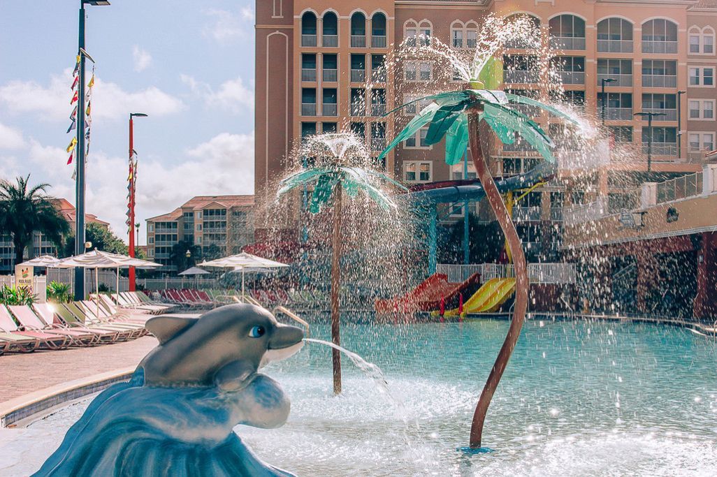 A swimming pool with a dolphin sculpture, water fountains, and a building with balconies in the background.