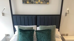 Een net opgemaakt bed met een blauwe deken en twee groene kussens, een witte handdoek op het bed, en een kunstwerk aan de muur erboven.