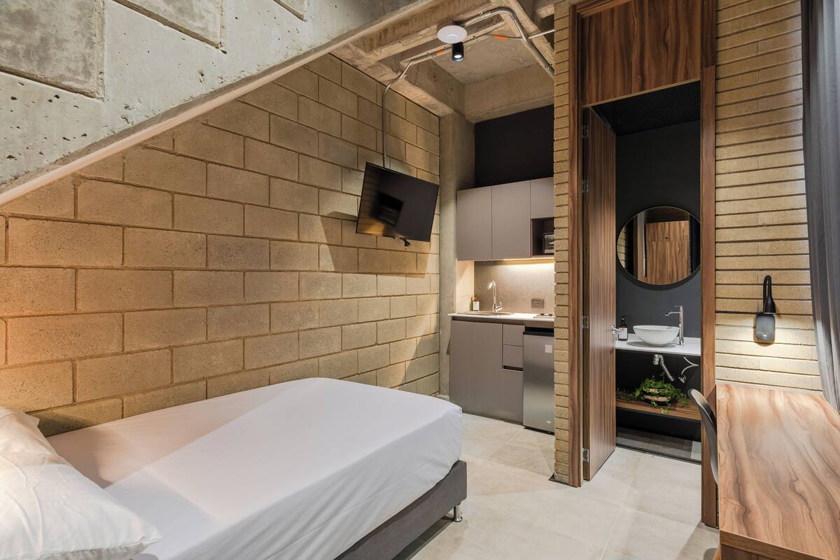 Une chambre moderne avec un mur en briques, un lit, un téléviseur fixé au mur, une porte en bois et un petit coin cuisine équipé d’un évier et d’un plan de travail.