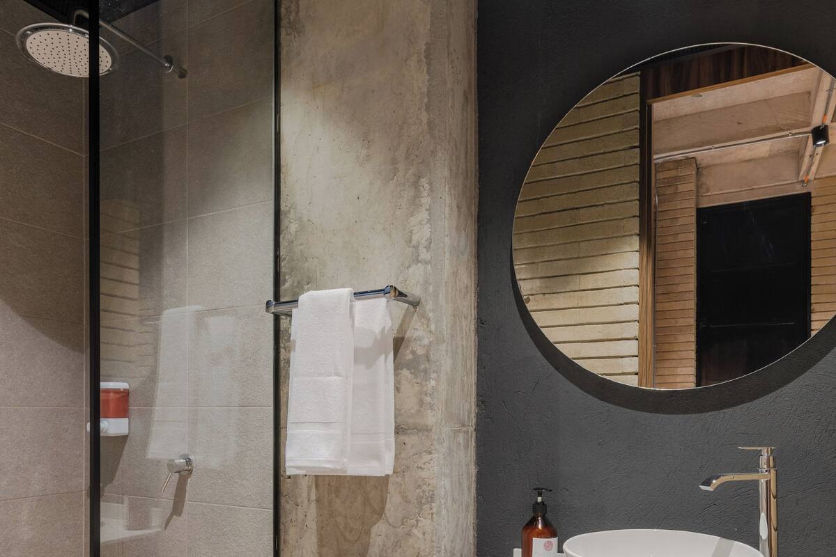 Une salle de bain moderne avec une douche à l’italienne en verre, un miroir rond, un lavabo blanc et une étagère en bois agrémentée d’une plante.