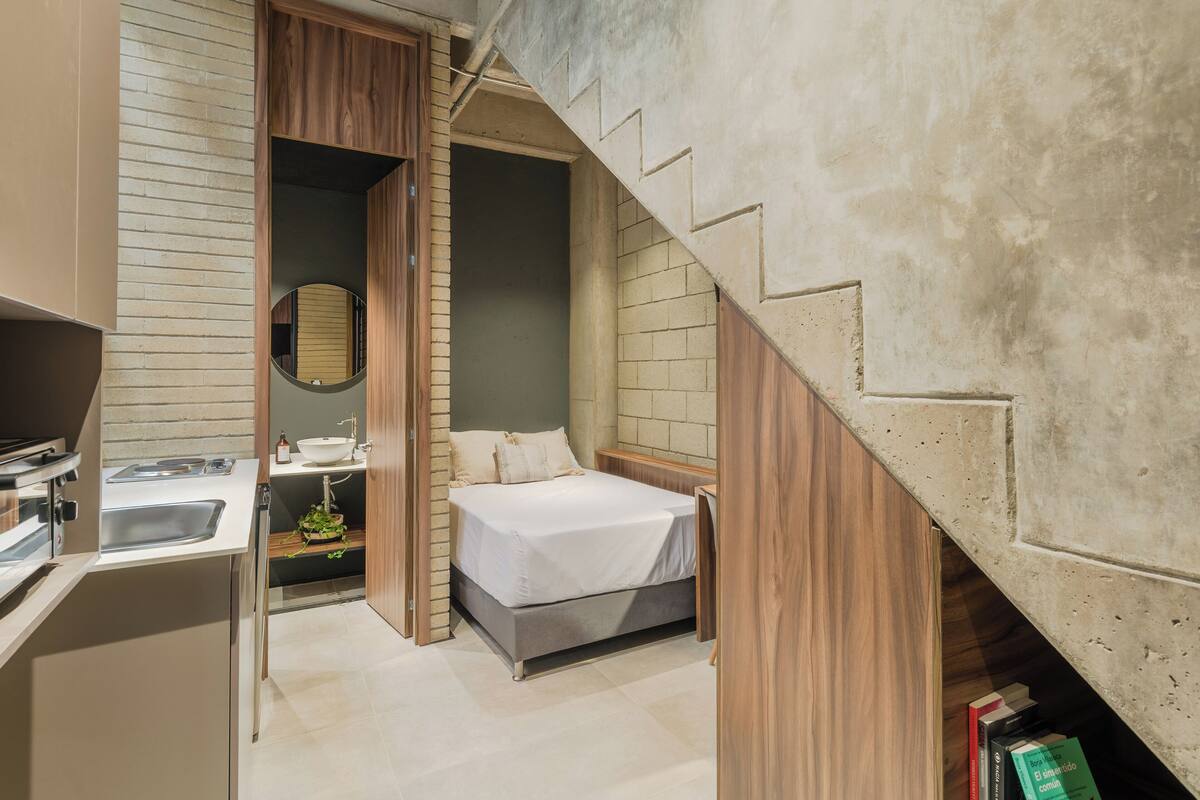 Une chambre à coucher de taille réduite, comprenant un lit, une table de chevet et une petite salle de bain avec un lavabo et un miroir.