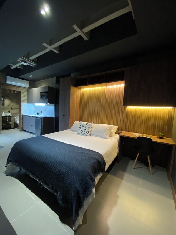 A modern hotel room with a large bed, bedside tables, a desk, and a built-in kitchen area.