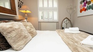 A bedroom with a large bed, fluffy pillows, a bedside lamp, a radiator, and a window with shutters.