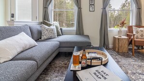 A cozy living room with a grey sofa, a coffee table with a beer bottle and glass, and a tray with snacks. - Cozy Acadia Hideaway with Hot Tub (Tremont)
