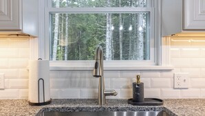 A kitchen with a stainless steel sink, a window with a view of trees, and white tiled walls. - Cozy Acadia Hideaway with Hot Tub (Tremont)
