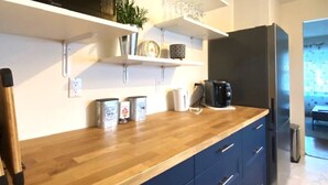 Une cuisine moderne avec des plans de travail en bois, un four à micro-ondes encastré et un réfrigérateur en acier inoxydable. Des étagères blanches sont fixées au-dessus du plan de travail et accueillent divers ustensiles de cuisine ainsi qu’une plante en pot.