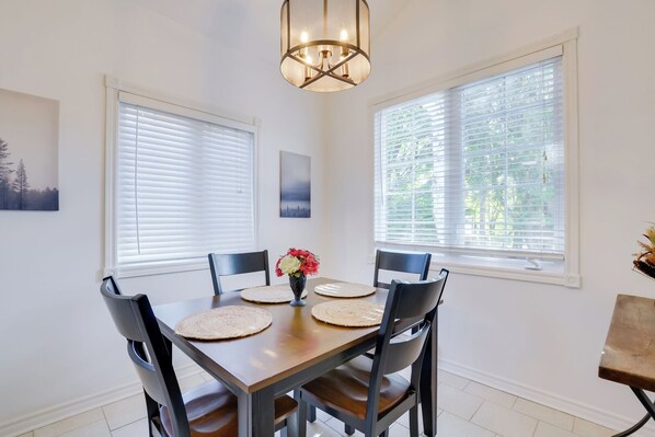 A dining area with a wooden table, four chairs, two windows with blinds, a pendant light, and a vase with flowers. - The little refuge (Gatineau)