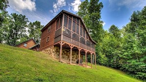 A large wooden house with a balcony, situated on a grassy hill with trees in the background.