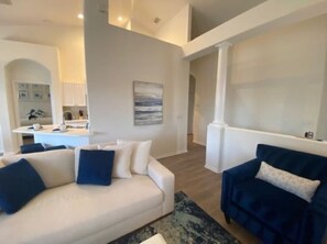 Uma sala de estar ampla, com um sofá branco e um sofá azul escuro, uma cozinha ao fundo e um quadro grande na parede.