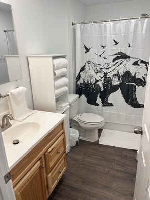 Ein Badezimmer mit weißem Waschbecken, Holz-Waschtisch, weißer Toilette und einer Duschvorhang mit Bergmotiv in Schwarz und Weiß.