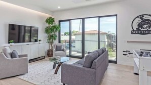 Una sala de estar moderna con un sofá, sillones, una mesa de centro de vidrio, un televisor y una mesa de futbolín.