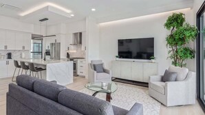Una cocina moderna con armarios blancos, una isla de mármol y un comedor con sillas. Hay un sofá gris y un sillón blanco en una sala de estar con un televisor grande y una planta.