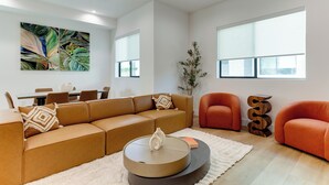 Ein modernes Wohnzimmer mit einem braunen Ecksofa, einem runden Couchtisch und einem weißen Teppich. Es gibt einen Essbereich mit Tisch und Stühlen, und ein großes Fenster sorgt für natürliches Licht.