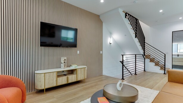 Ein modernes Wohnzimmer mit einem Flachbildfernseher an einer Holzwand, einem Holz-Konsolentisch, einem runden Couchtisch mit einem Buch, einer Treppe mit schwarzer Geländer und einem Sofa.