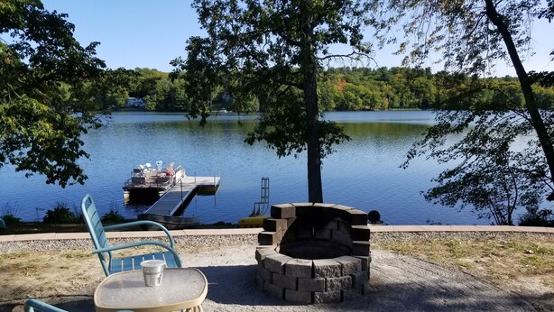 A lakeside patio with a fire pit and two chairs, overlooking a calm lake with a dock and boat. - Blue Loon Camp on Pleasant Pond in Litchfield (Litchfield)