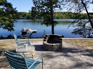 A lakeside patio with a fire pit and two chairs, overlooking a calm lake with a dock and boat. - Blue Loon Camp on Pleasant Pond in Litchfield (Litchfield)