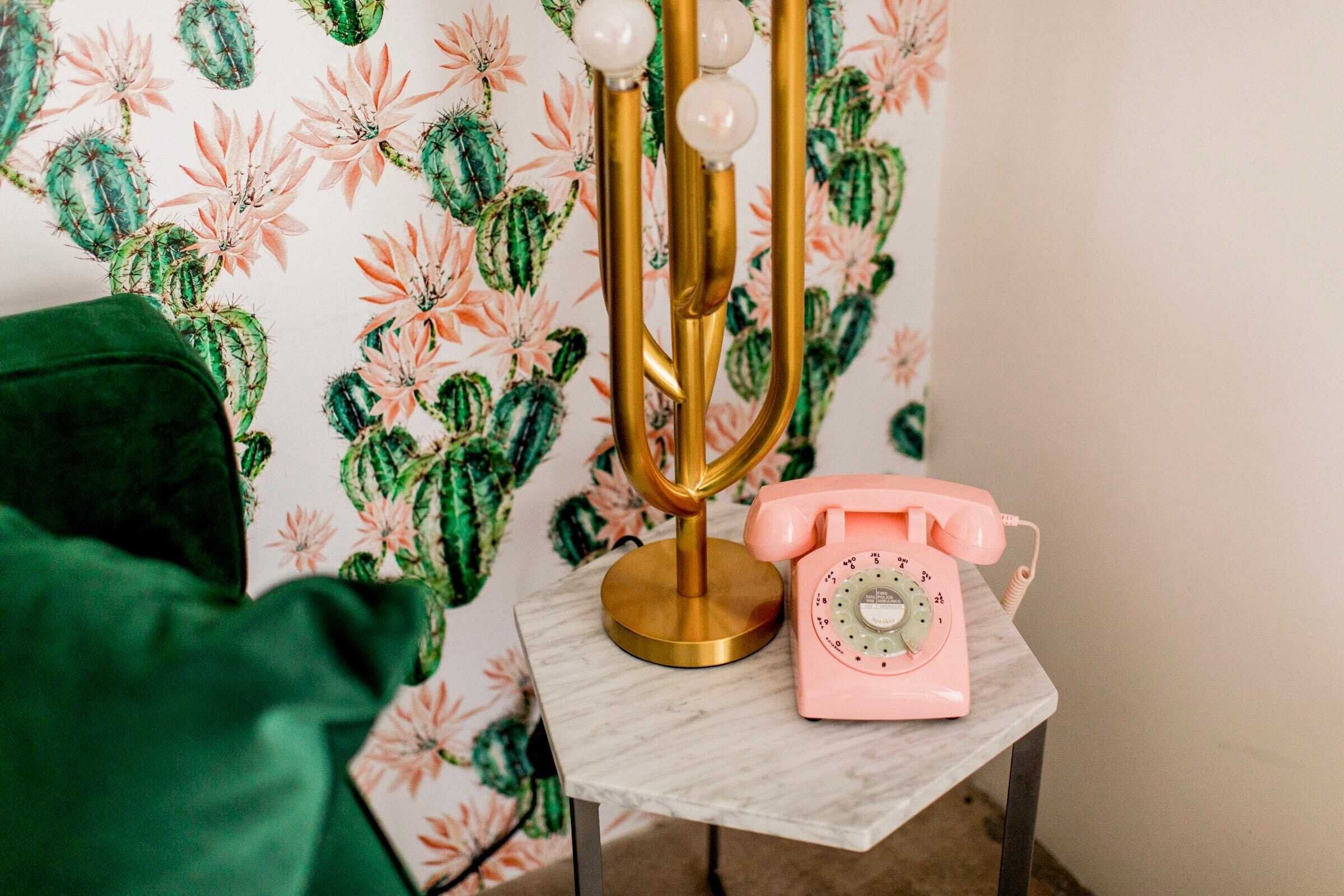 Ein kleiner Tisch mit einer Goldlampe und einem rosafarbenen Wählscheibentelefon, vor einer Tapete mit Kakteen- und Blumenmustern.