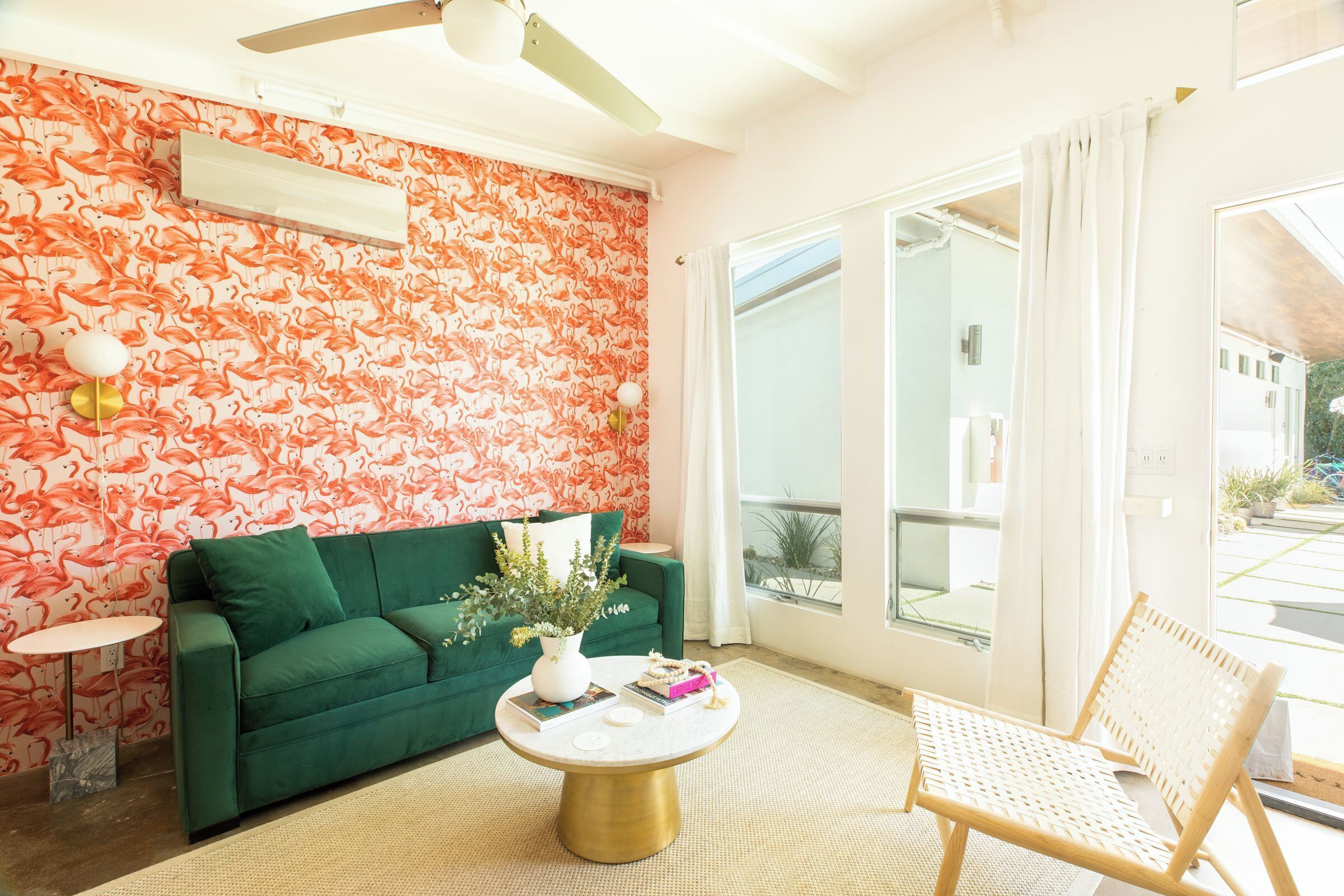 Une pièce avec un canapé vert, une table basse ronde, un fauteuil en osier, un vase blanc contenant des plantes et un mur orné d’un motif de flamants roses.