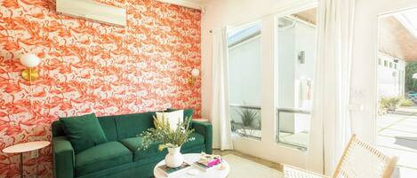 A living room with a green sofa, a round coffee table, and a chair. There is a window with curtains and a view of the outside.