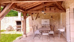 A covered outdoor patio with a stone wall, a wooden ceiling, a table with chairs, and a built-in oven.