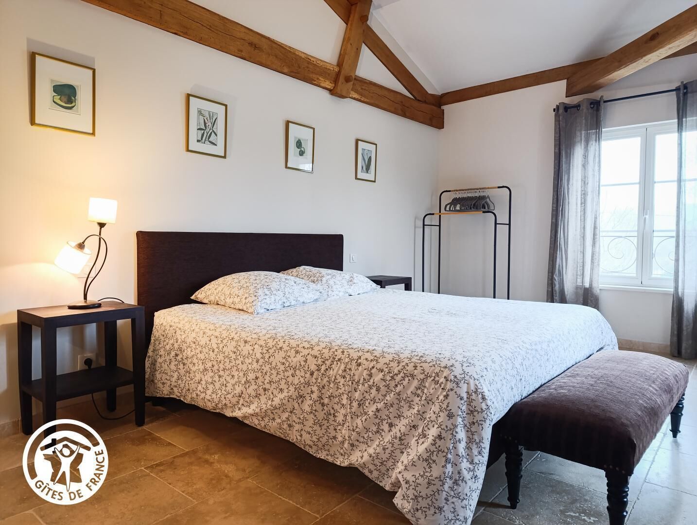 Une chambre à coucher avec un lit, une table de chevet avec une lampe, un banc, un portant à vêtements et des œuvres d’art encadrées accrochées au mur.